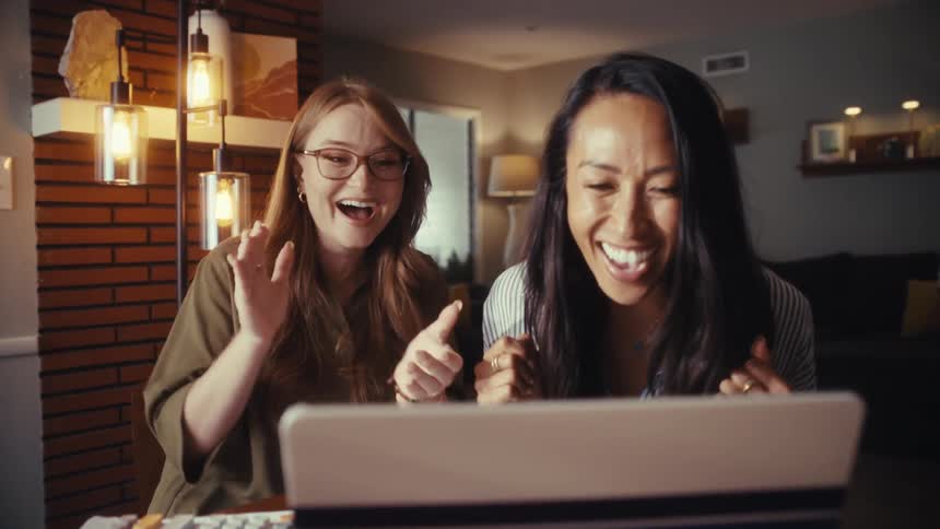A slow push-in shot shows two excited women celebrating good news they received on their laptop while working from home