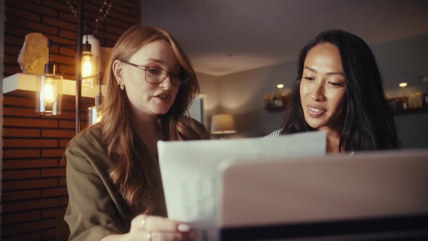 Two diverse female colleagues review and discuss charts on a document while working from home in slow motion