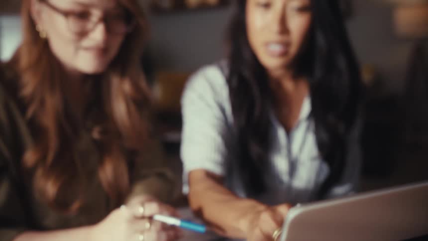 Two diverse professional women collaborate on a project, reviewing data on a laptop and charts in a home office