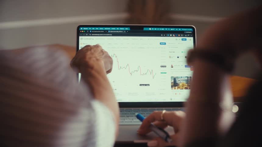 An over-the-shoulder shot shows two people analyzing a fluctuating financial graph on a laptop screen