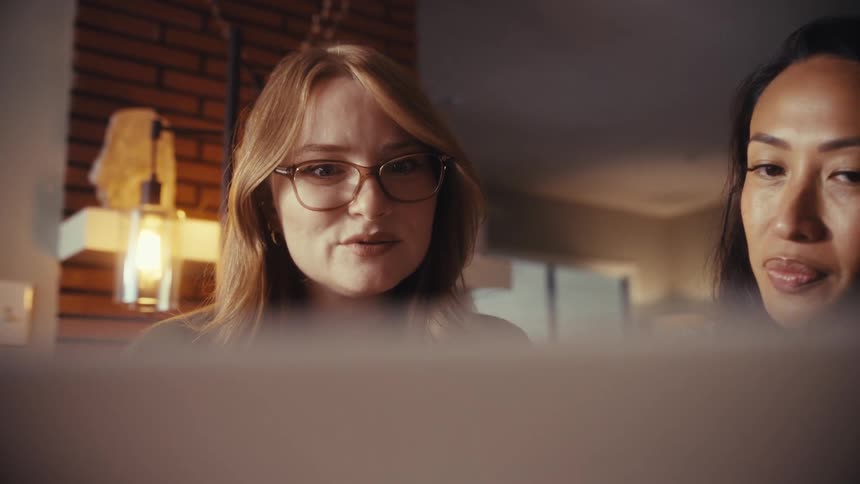 Two diverse female colleagues work together, discussing a project on a laptop in a warmly lit, modern home office