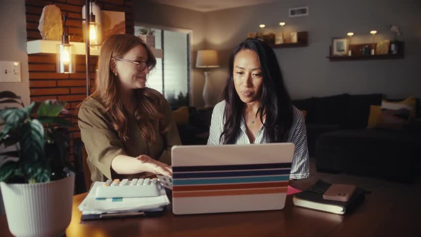 Two female colleagues work on a project at home, using a laptop and calculator, and celebrate their success with a happy handshake