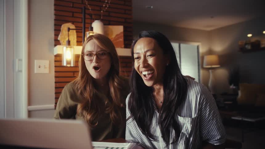 Two diverse and happy women celebrate receiving good news on their laptop, laughing and cheering together in a sunlit room