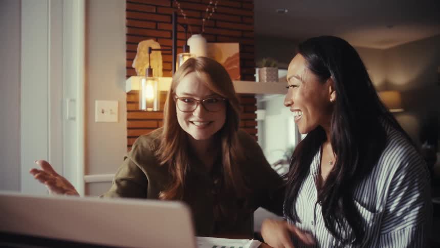 Excited female colleagues laugh and cheer at good news on their computer while working together from home