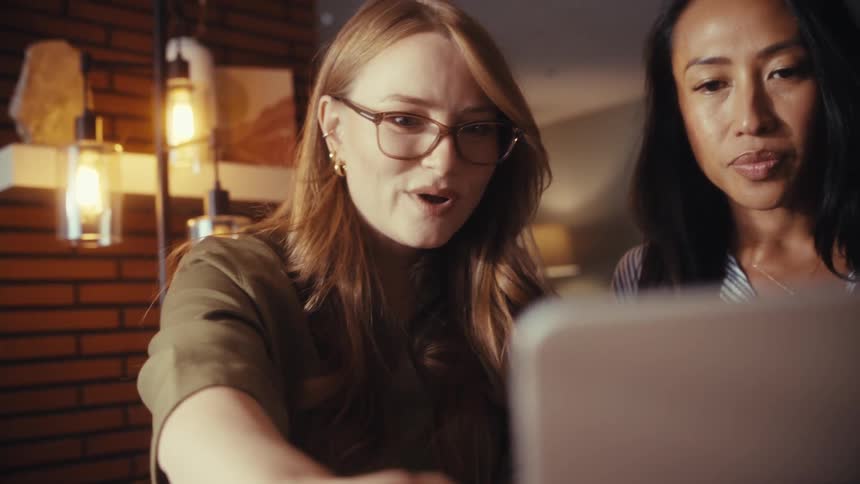 Two diverse female colleagues collaborate on a project, pointing at a laptop screen and discussing their work in a modern office