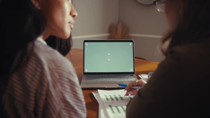 Over the shoulder view of two diverse women collaborating on a project and pointing at a laptop with a green screen on a desk with financial documents