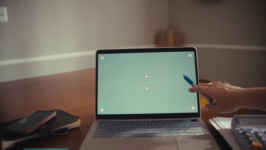 Two women collaborate at a desk as one points to a laptop's green screen with tracking markers, viewed from over their shoulders