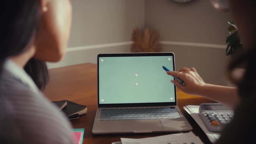 A woman points to a green screen on a laptop while collaborating with her coworker at a desk