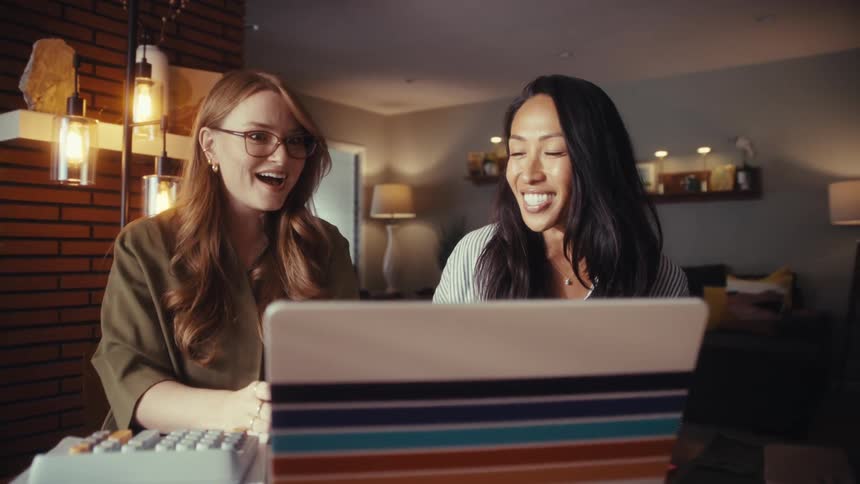 Two diverse female entrepreneurs smile and discuss their project on a laptop while working together from a cozy home office