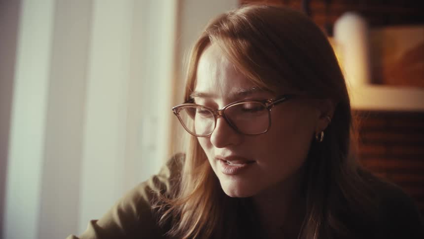 Close-up of a friendly young businesswoman with red hair and glasses talking and smiling during a video conference in a warmly lit room