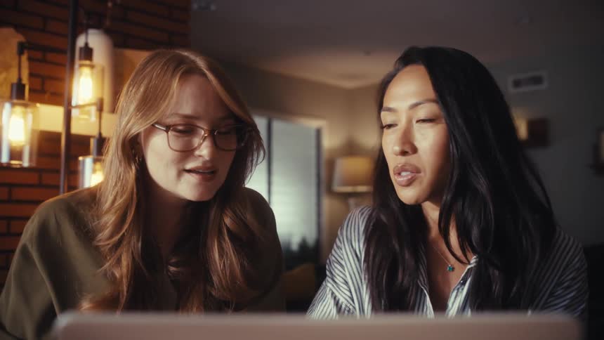 Two professional women collaborate on a project from a home office, pointing at the laptop screen as they discuss their work in the evening