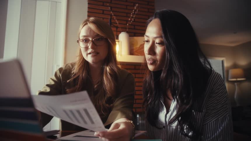 Two professional women collaborate while reviewing a business report with charts and data on a laptop in a cozy home office