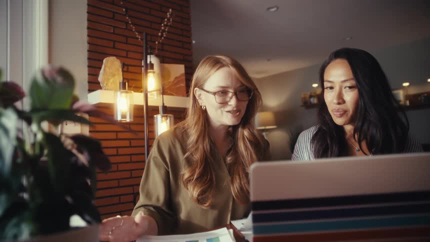 Two diverse professional women happily collaborate on a project using a laptop and charts in a modern home office