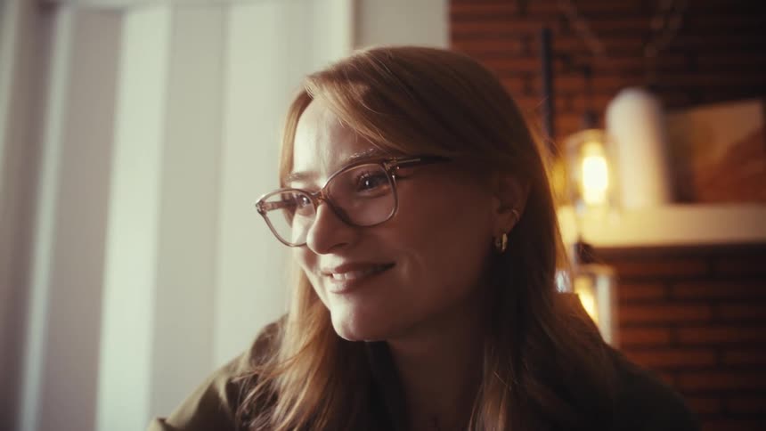 A close-up shot of a smiling young woman with red hair and glasses looking thoughtfully at her screen