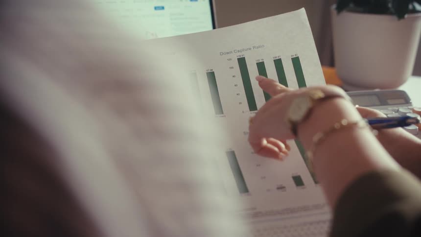 Over the shoulder shot of a financial advisor explaining market statistics on a printed document with a laptop in the background