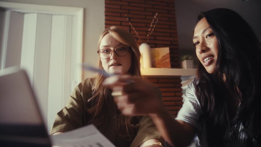 Two diverse female colleagues collaborate in a warmly lit office, pointing at a laptop screen while analyzing data charts