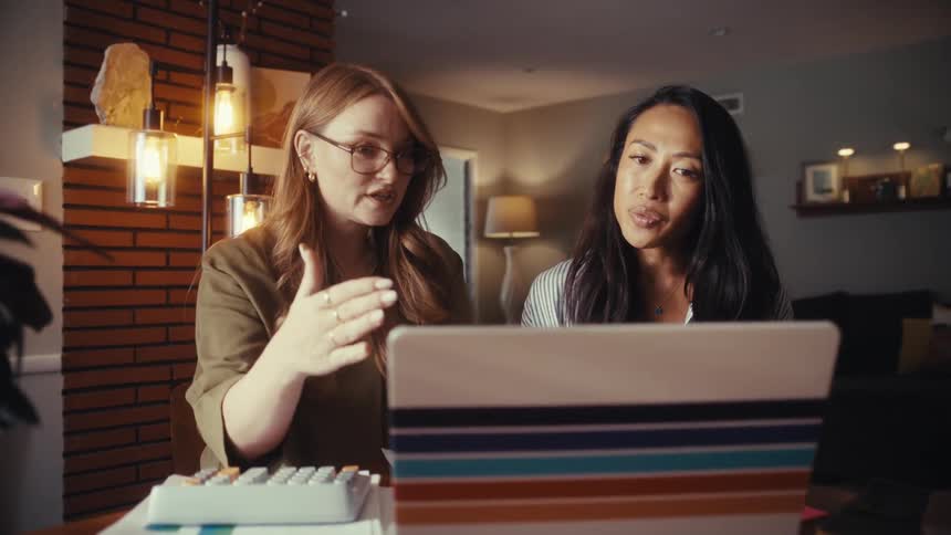A focused woman with glasses explains information on a laptop to her colleague as they collaborate on a project from home
