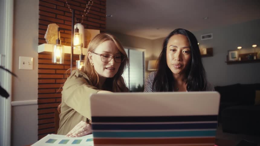 Two diverse female colleagues collaborate on a project, pointing at a laptop screen while working from a modern home office