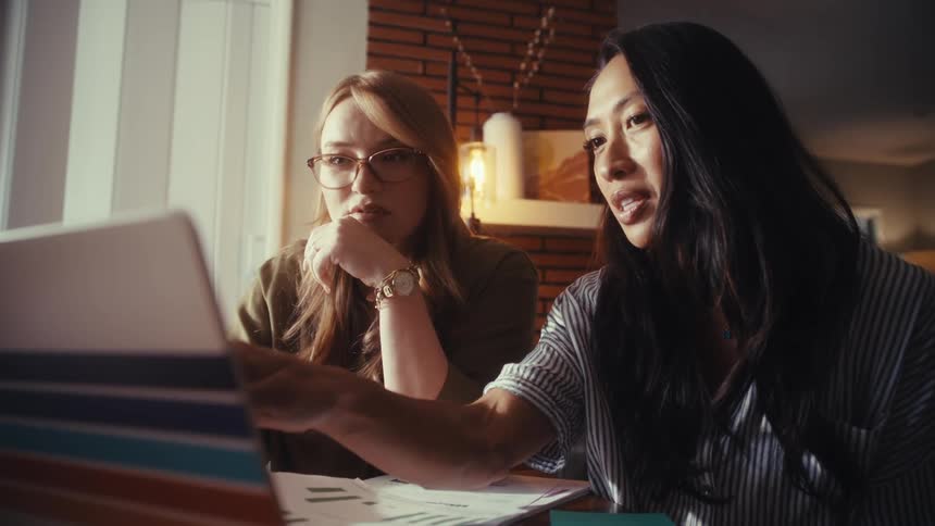 Two diverse women work together on a laptop, pointing at the screen and discussing a project in a home office setting