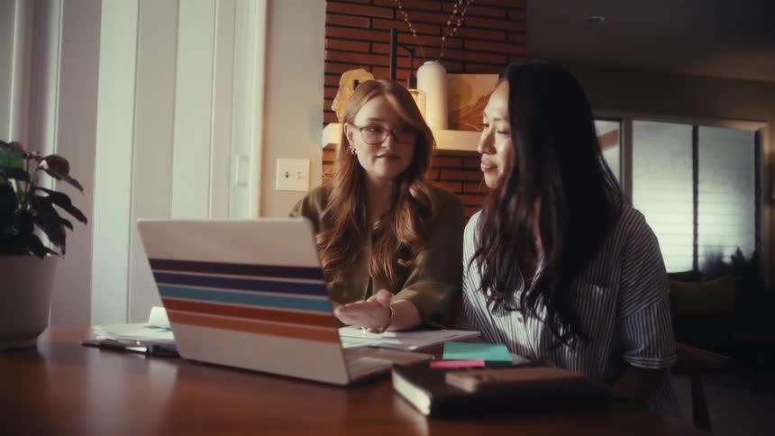 Two professional young women work together and collaborate on a project using a laptop in a modern home office