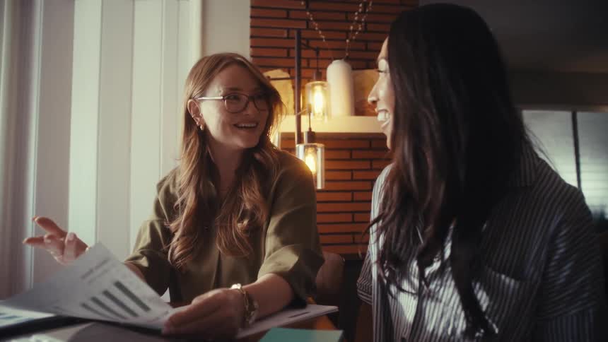 Two diverse businesswomen happily collaborate on a project, analyzing charts on paper and a laptop in a modern home office