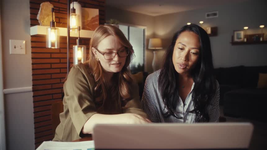 Two diverse female colleagues work together, discussing a project on a laptop in a modern home office setting