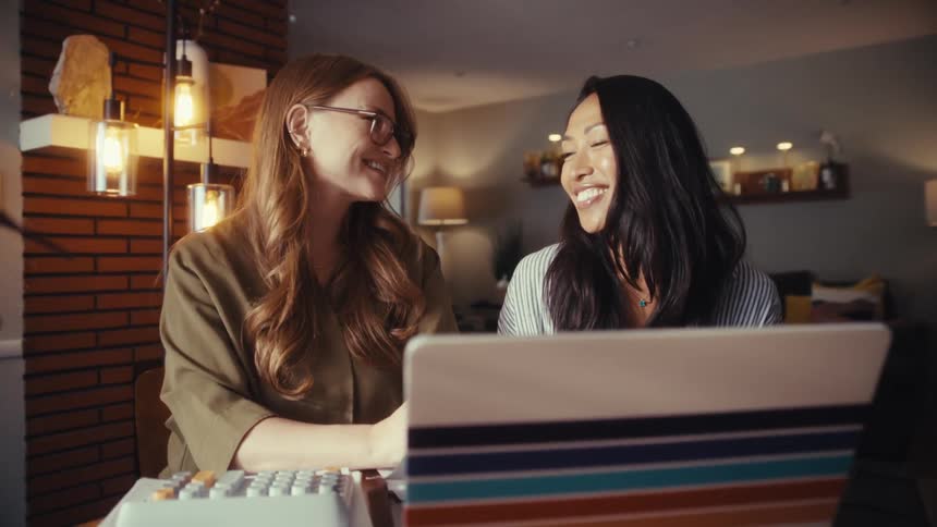 Two cheerful female colleagues collaborate and smile while looking at a laptop screen in a modern home office