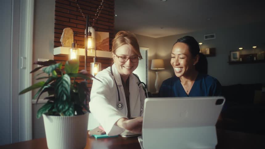 A smiling doctor and her nurse colleague happily collaborate on a project using a tablet in a warmly lit home office