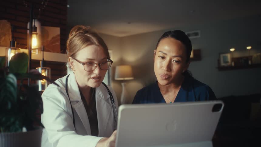 A female doctor and nurse collaborate late at night, reviewing information on a digital tablet in a cozy home office