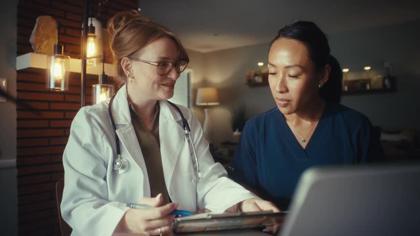 A female doctor and nurse collaborate, discussing notes on a clipboard and smiling while working together in a warmly lit office