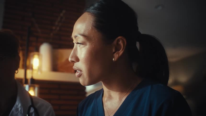 A female nurse in blue scrubs seriously discusses a case with a fellow female doctor in a dimly lit room