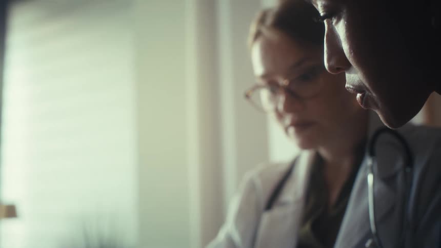 A close-up rack focus shot shows two diverse female doctors collaborating on a case file in a modern, sunlit office
