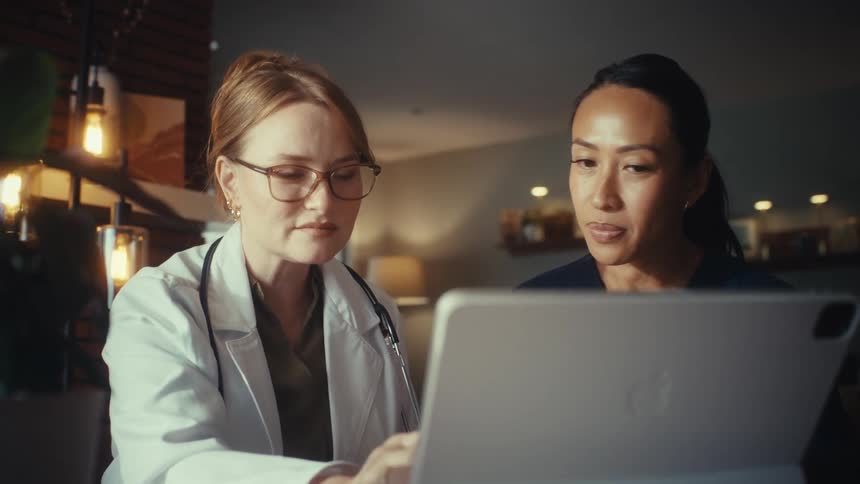 A female doctor and nurse collaborate while reviewing information together on a tablet in a warmly lit office