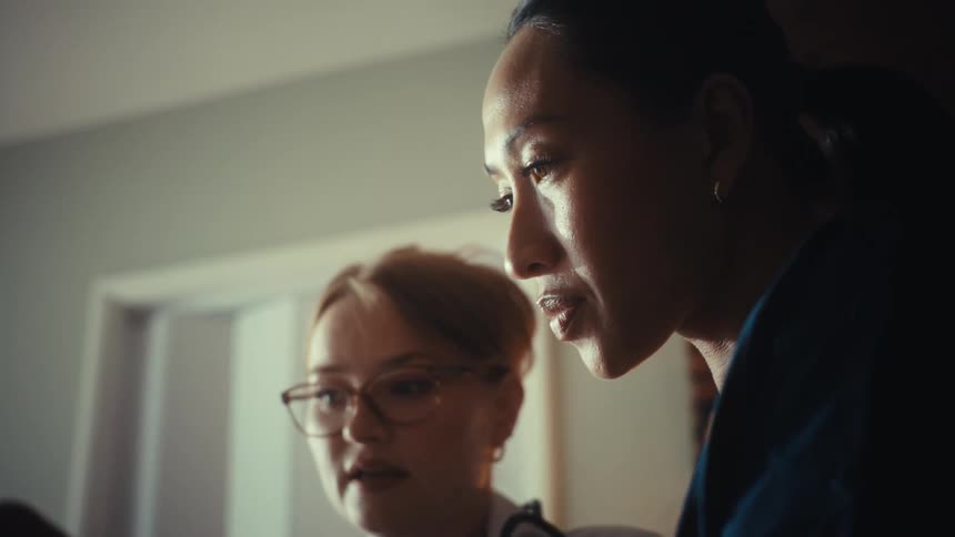 A low-angle close-up of a female doctor with glasses and a nurse seriously reviewing medical data together in a dimly lit office