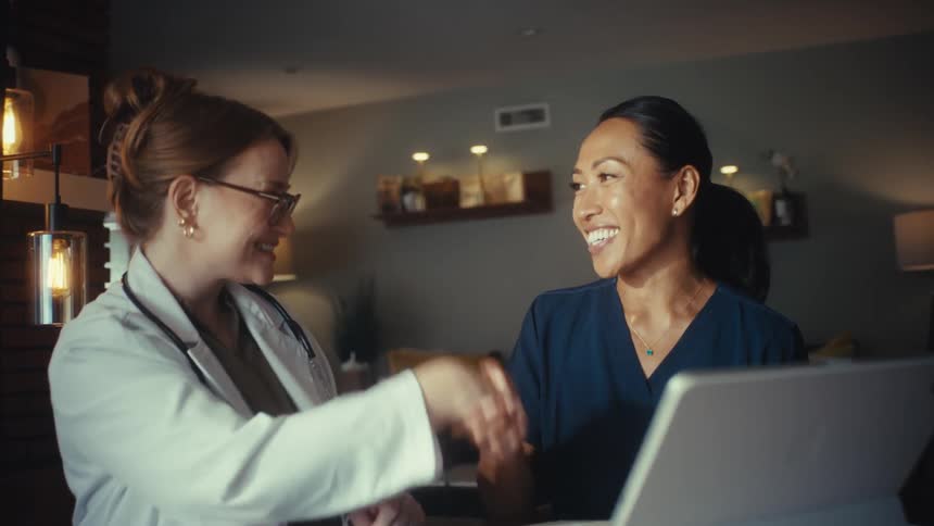 A female doctor in a white coat and a nurse in scrubs happily shake hands after a successful collaboration using a tablet in a modern office
