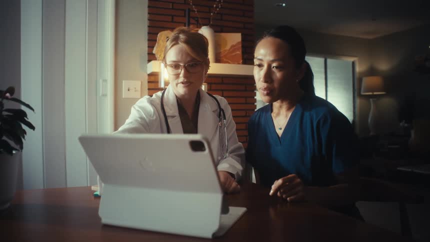 Two healthcare workers, a doctor and a nurse, consult together while looking at a tablet at a dining room table