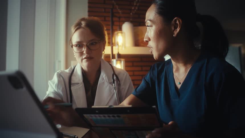 A dedicated female doctor and nurse collaborate late at night, reviewing medical charts on a tablet and smiling together