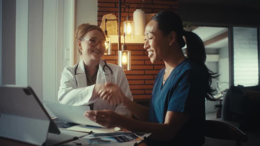 A female doctor and nurse collaborate while reviewing patient files, smiling and shaking hands in agreement