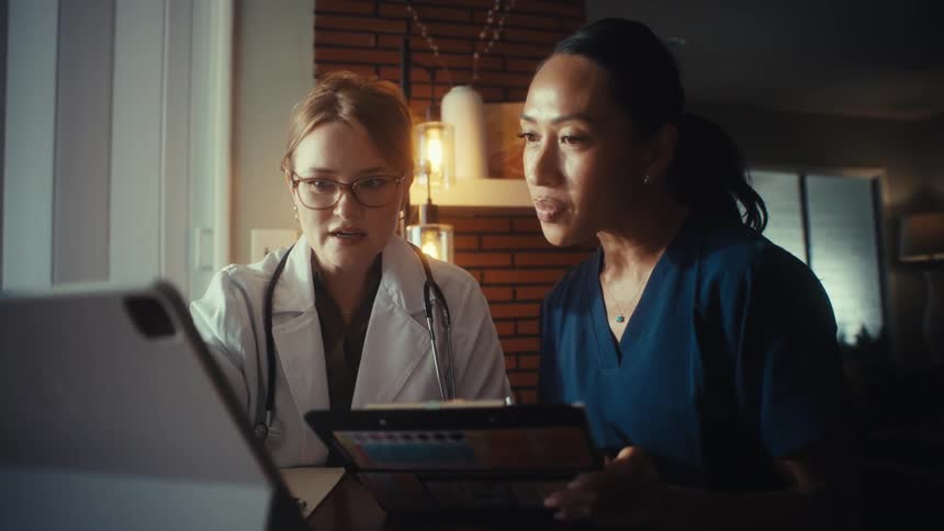A female doctor and nurse collaborate while reviewing patient records on a digital tablet in a dimly lit home office