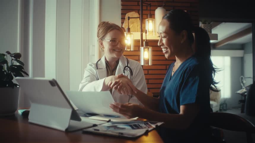 Two smiling female medical professionals review documents and shake hands to celebrate a successful collaboration in a warmly lit office