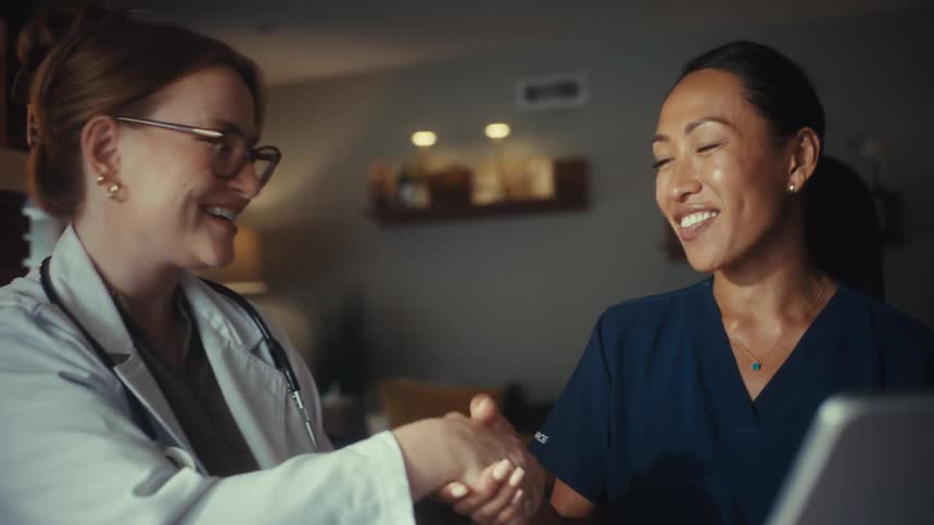 Two smiling female medical professionals, a doctor and a nurse, review information on a tablet before shaking hands in a successful collaboration