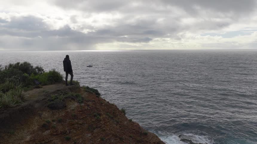 A solitary figure in a hooded jacket stands on a windswept cliff, gazing out at the vast ocean under a moody, overcast sky