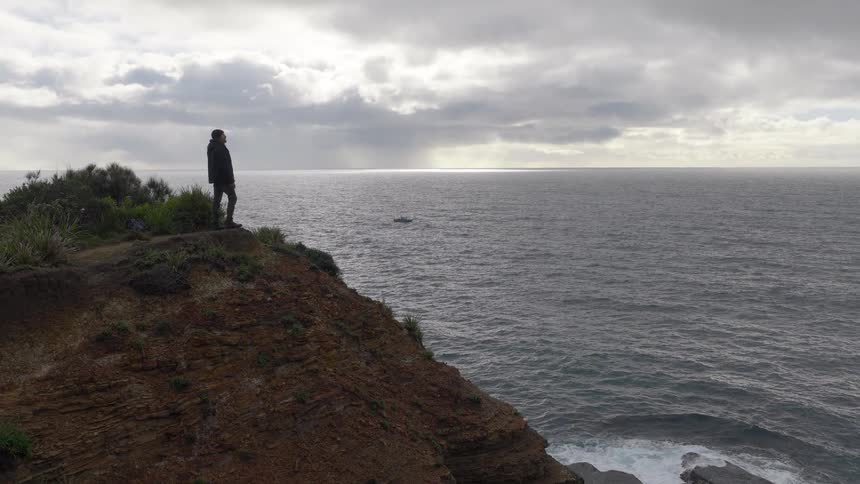 A lone figure stands on a dramatic cliffside, contemplating the vast, restless ocean under a cloudy sky as the camera slowly pulls back