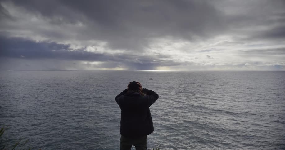 A lone person stands on a cliff overlooking a vast ocean under a dramatic, cloudy sky, pulling up their hood in slow motion