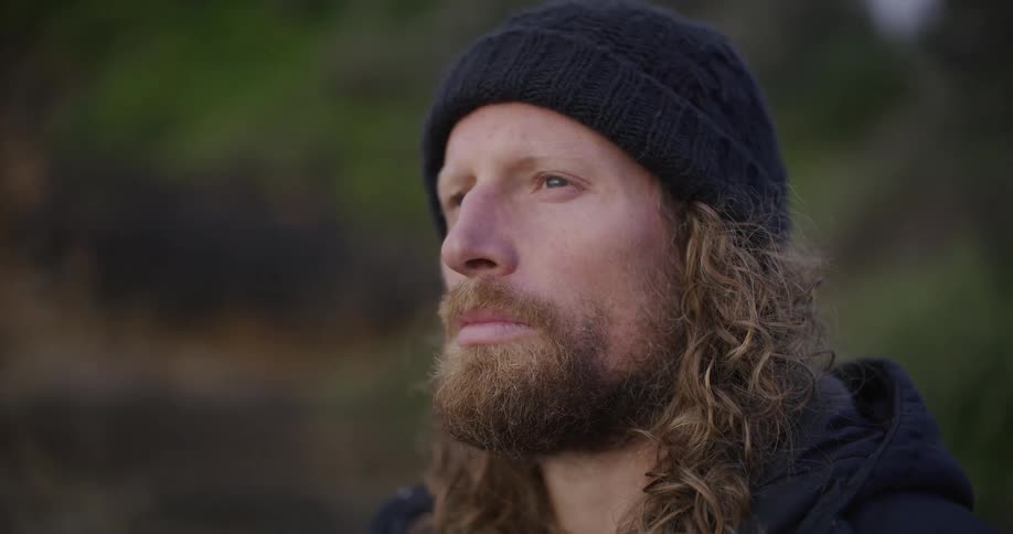 Close-up profile of a contemplative man with long curly hair and a beard, wearing a black beanie and looking into the distance