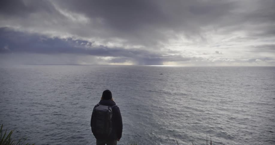 A lone hiker with a backpack gazes out at the vast ocean and a distant boat under a dramatic, stormy sky