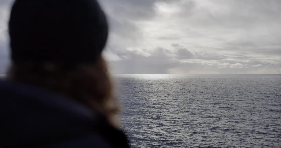 A bearded man in a beanie gazes thoughtfully at the sunlit horizon across the vast, calm ocean from the deck of a boat