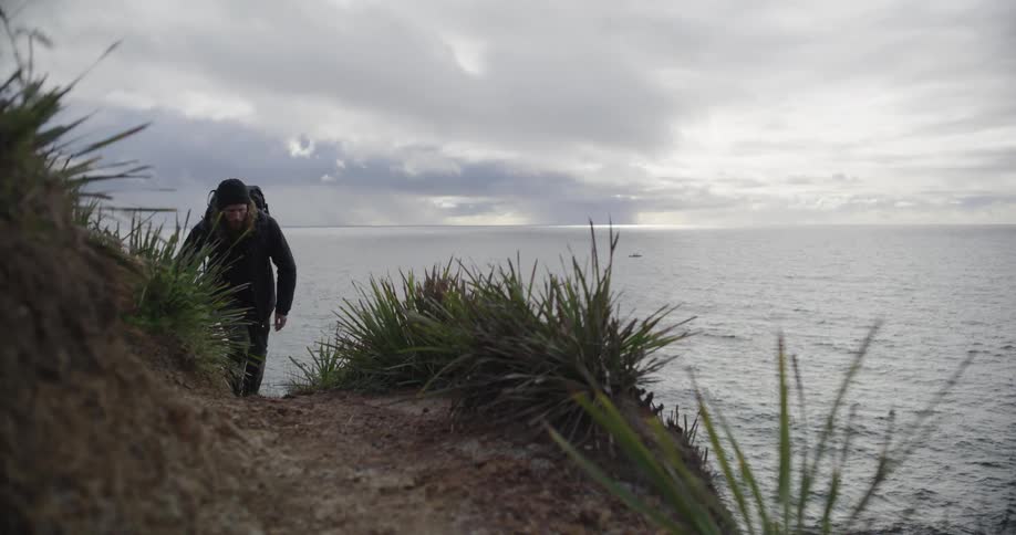 A rugged hiker with long hair and a backpack carefully makes his way up a steep coastal cliff path on an overcast day
