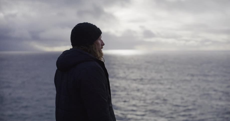 A pensive man in a winter jacket and beanie contemplates the vast, moody ocean on an overcast day