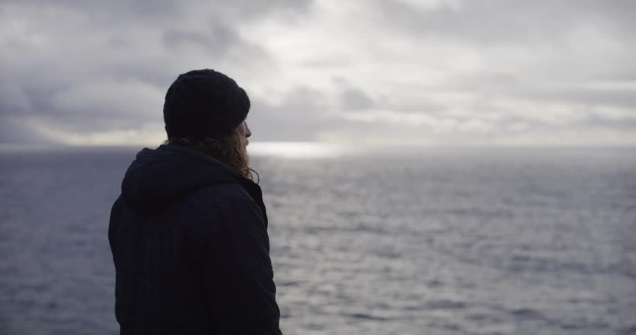 A pensive man with long hair wearing a beanie and jacket looks out at the vast, moody ocean under an overcast sky